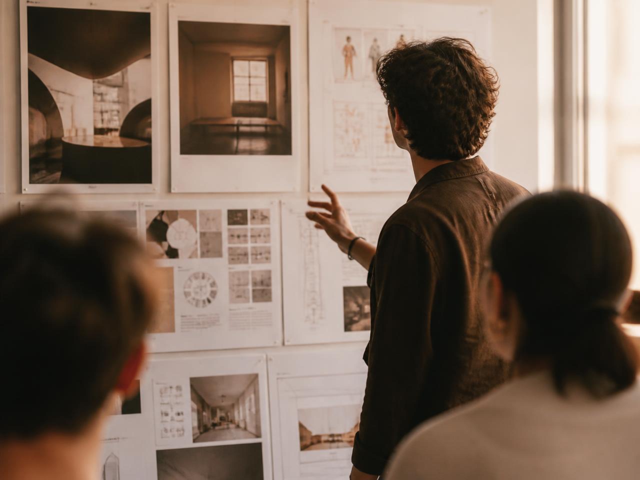 A young adult presenting a wall of pinned portfolio work to two people