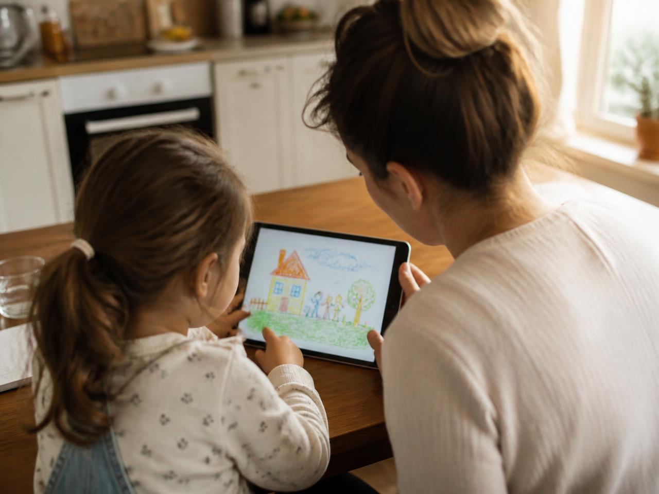 A parent and child at a kitchen table looking together at a drawing on a tablet